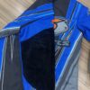 A SLACK Driving Jacket in blue and gray with a black patch on the sleeve and a logo design featuring an abstract bird on the chest, laid flat on a wooden surface.