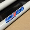 Close-up of a metal frame tube with the words "Foot Pan - For Elevate, Pursuit & Xpect CHAMP KART" in stylized blue and red letters, against a background of black and tan surfaces.