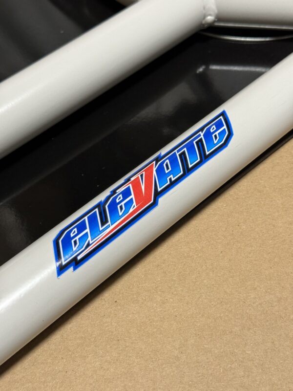 Close-up of a metal frame tube with the words "Foot Pan - For Elevate, Pursuit & Xpect CHAMP KART" in stylized blue and red letters, against a background of black and tan surfaces.
