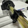 Close-up of a mechanical assembly featuring the Foot Pan for Elevate, Pursuit & Xpect CHAMP KART, with a cylindrical metal rod held in place by clamps and mounted on a white structure.