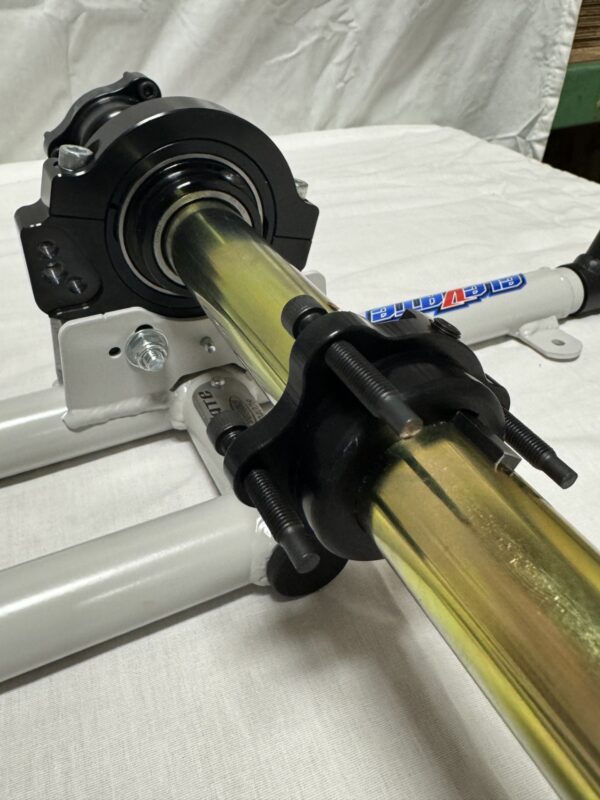 Close-up of a mechanical assembly featuring the Foot Pan for Elevate, Pursuit & Xpect CHAMP KART, with a cylindrical metal rod held in place by clamps and mounted on a white structure.