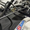 Close-up of a mechanical assembly featuring metal rods, a bracket, various fasteners, and a cable. The frame has a "Foot Pan - For Elevate, Pursuit & Xpect CHAMP KART" logo on it.