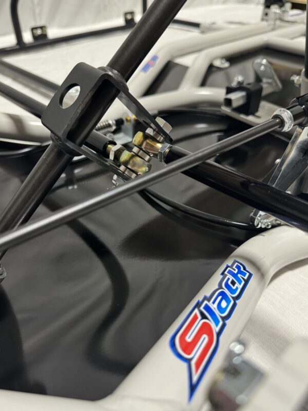Close-up of a mechanical assembly featuring metal rods, a bracket, various fasteners, and a cable. The frame has a "Foot Pan - For Elevate, Pursuit & Xpect CHAMP KART" logo on it.