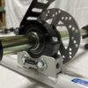 Close-up of a mechanical assembly featuring a Foot Pan from Elevate, Pursuit & Xpect CHAMP KART, showcasing a metal rod, a bearing with sleek black housing, and a perforated disc, all mounted on a pristine white framework.