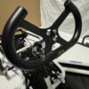 Close-up of a black steering wheel attached to an incomplete frame setup, likely for a small vehicle or simulator. Background consists of a beige and white fabric backdrop, featuring the Foot Pan designed for Elevate, Pursuit & Xpect CHAMP KART.