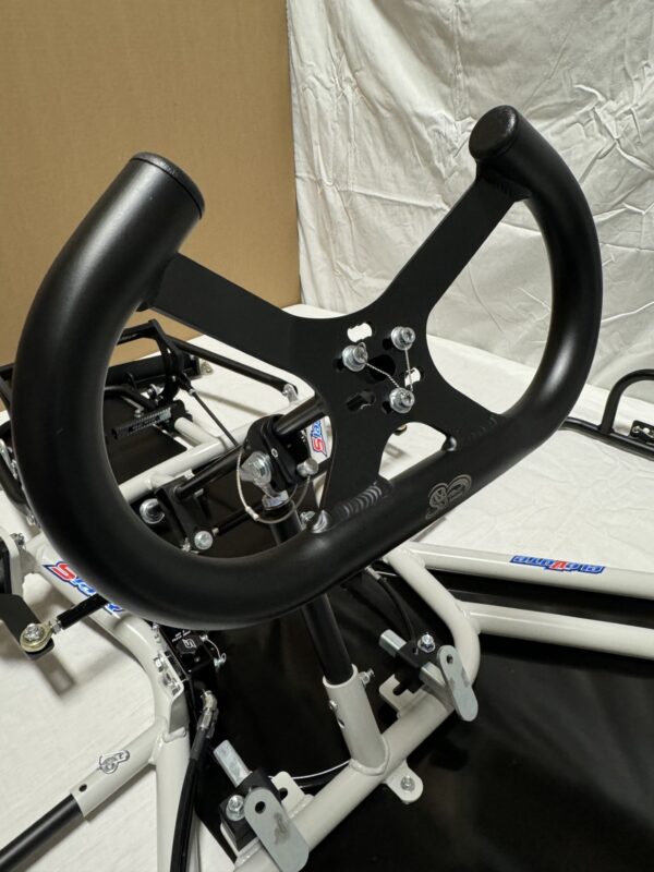 Close-up of a black steering wheel attached to an incomplete frame setup, likely for a small vehicle or simulator. Background consists of a beige and white fabric backdrop, featuring the Foot Pan designed for Elevate, Pursuit & Xpect CHAMP KART.