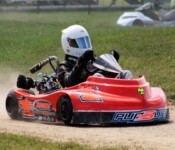 A person in a racing suit and helmet drives a red go-kart on a dirt track, kicking up dust. Another go-kart can be seen blurred in the background.