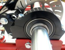 Close-up view of a mechanical component featuring a cylindrical metal shaft, a bearing, bolts, and part of a brake disc. The assembly appears to be part of a machine or vehicle.
