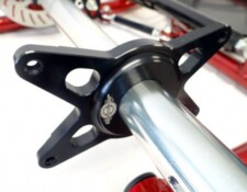 Close-up image of a metallic component with a black mount attached, possibly part of a mechanical or automotive assembly. Red parts are visible in the background.