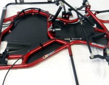 A close-up view of a red metal go-kart frame with a black seat panel, various mounting brackets, and steering components on a white background.